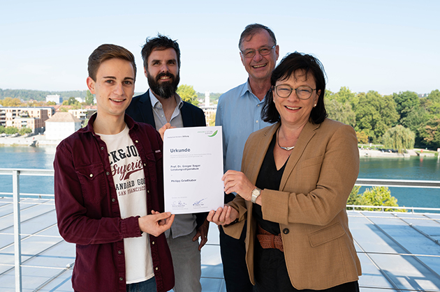 Das Bild zeigt den Preisträger sowie die Stifter Herrn Dr. Tögel, Frau Prof. Dr. Rein und Prof. Dr. Thomas Birkhölzer. Sie stehen auf einer Dachterrasse und zeigen stolz die Urkunde des Stipendiaten.