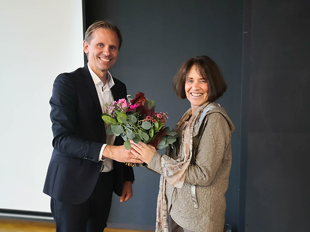 Das Bild zeigt Gunnar Schubert und Annette Kleinfeld vor einer Tafel. Sie sind schick gekleidet und lächeln in die Kamera. Schubert übergibt Kleinfeld einen Blumenstrauß.