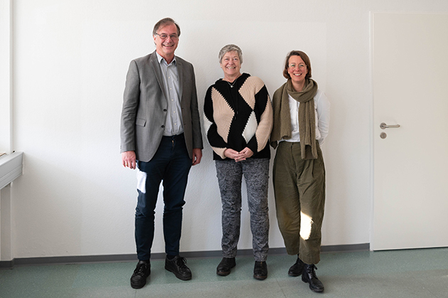 Gruppenfoto vor einer weißen Wand, von links nach rechts: Thomas Birkhölzer trägt eine schwarze Jeans, graues Hemd und Jackett. In der Hand hält er den Kooperationsvertrag. In der Mitte steht Hanna Schönfeld, sie trägt eine graugemusterte Hose und einen schwarz-beige gemusterten Strickpullover, rechts steht Sandra Flügel in khakifarbener Hose und Schal und weißem Shirt.