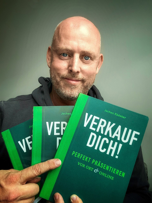 Jochen Rädeker trägt einen dunklen Kapuzenpullover, lächelt und zeigt drei Exemplare seins Buchs in der Hand