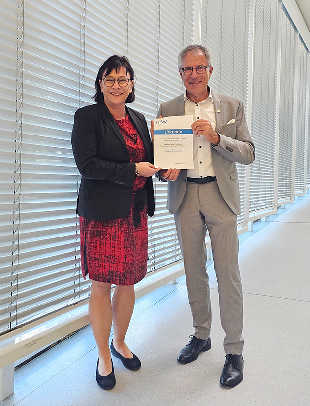 HTWG News_Beitritt HfSW Das Bild zeigt Prof. Wolfmaier und Prof. Rein in einem grauen Gang. Sie halten gemeinsam die Urkunde zum Beitritt der HTWG in die HfSW