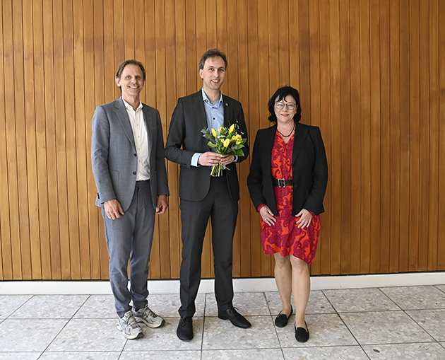 Zwei Männer und eine Frau stehen bei einer Gratulation vor einer Wand