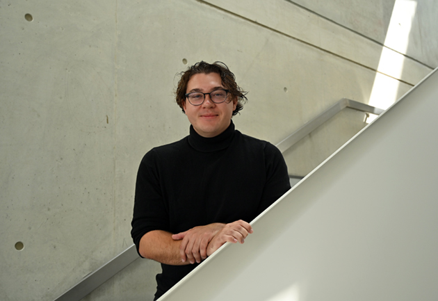 Ein junger Mann mit locken und Brille lehnt an einem Treppenaufgang