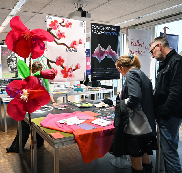 Zwei Personen schauen sich bunt gedruckte Karten an, die auf einem Tisch ausliegen. Im Hintergrund hängen bunte Poster von der Decke
