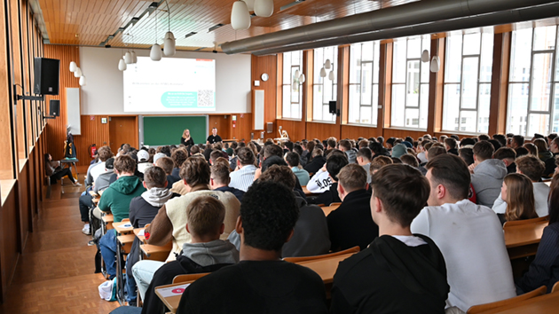 Das Foto zeigt von hinten eine große Gruppe von Menschen sitzend in einem holzvertäfelten Hörsaal. Vorne stehen zwei Menschen und sprechen.