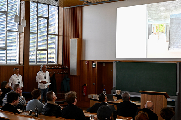 Zwei Männer stehen in einer Aula vor Publikum und schauen auf eine Präsentation