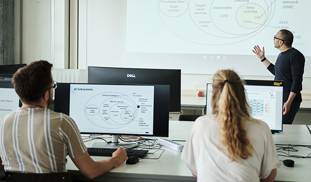 Ein Mann steht vor einer Präsentation, zwei Studierenden hören zu und schauen zu ihm