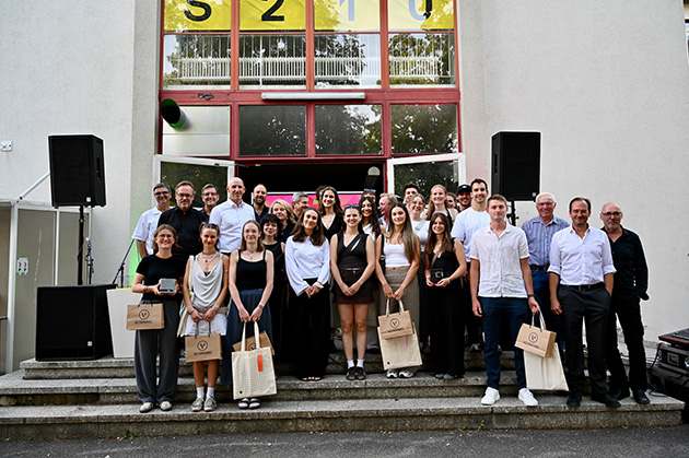 Große Gruppe von etwa 25 Personen steht auf den Außentreppen eines Gebäudes, mehrere junge Preisträgerinnen in der vorderen Reihe halten Auszeichnungen in der Hand. Im Hintergrund ist der Gebäudeeingang mit großen Fensterfronten zu sehen.