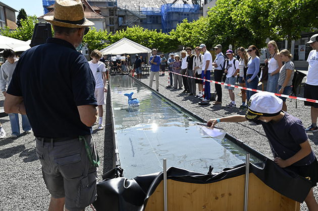 Menschen stehen an einem langen, schmalen Wasserbecken im Freien und beobachten ein kleines blaues, walförmiges Modellboot im Wasser. Im Vordergrund ein Mann mit Strohhut, rechts ein Junge mit Cap, der sich über das Becken beugt.