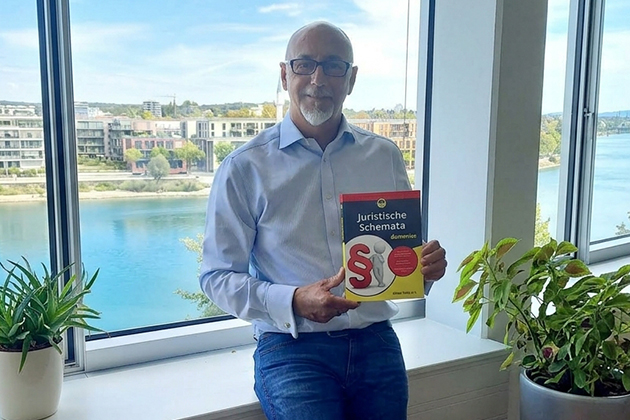 Ein Mann mit einem Buch mit dem Titel "Juristische Schemata für Dummies" lächelt in die Kamera. Im Hintergrund sieht man einen Fluss und Häuser.