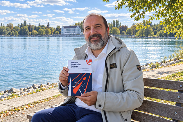 Ein Mann sitzt mit einem Buch in der Hand auf einer Bank, es ist herbstlich, im Hintergrund sieht man einen See