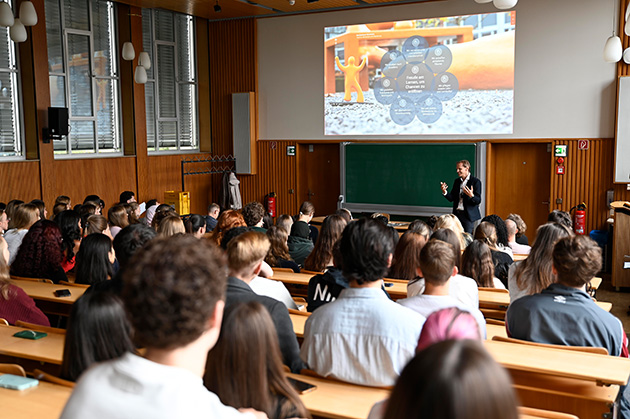 Hinterköpfe junger Menschen in einer Aula, vorne spricht ein Mann im Anzug.