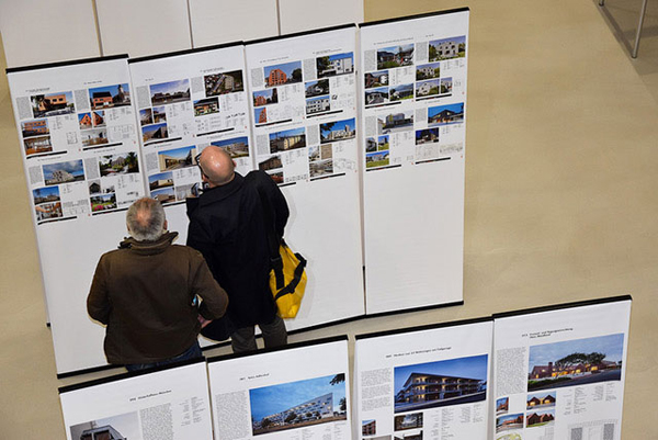 Ein Ausstellungsraum voller Stelen von oben fotografiert. Auf den Stelen stehen Informationen neben Bildern von Gebäuden. Zwei Männer stehen vor einer Stele und betrachten sie.