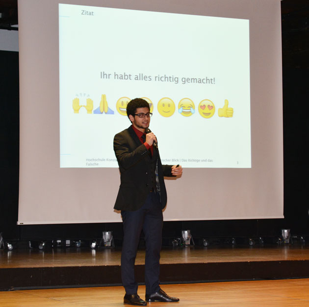 Ein junger Mann mit Mikro auf einer Bühne. Auf der Leinwand hinter ihm steht "Ihr habt alles richtig gemacht". Unter dem Satz ist eine Reihe von Emojis abgebildet.