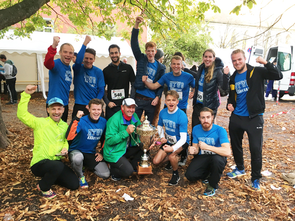 12 Menschen in Laufkleidung, teilweise mit Startnummern auf den T-Shirts, posieren mit Pokal für ein Gruppenfoto. Einige recken die Fäuste in die Luft.