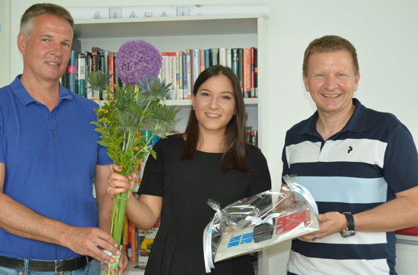 Eine junge Frau steht zwischen zwei Männern vor einem Bücheregal. Sie hält einen Blumenstrauss und ein Geschenk in der Hand.