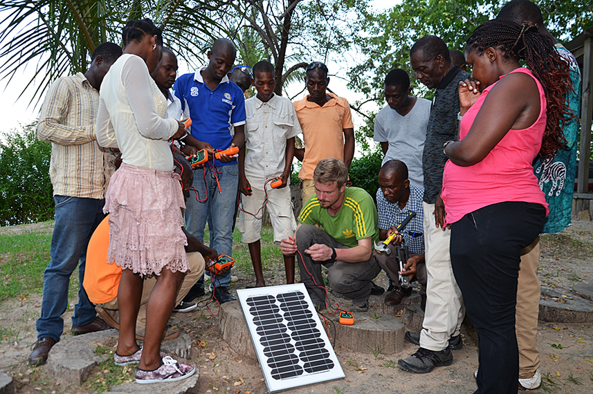 Eine Gruppe von Menschen steht im Kreis um ein Solarmodul. Zwei Männer knien hinter dem Modul und arbeiten an einem Kabel.
