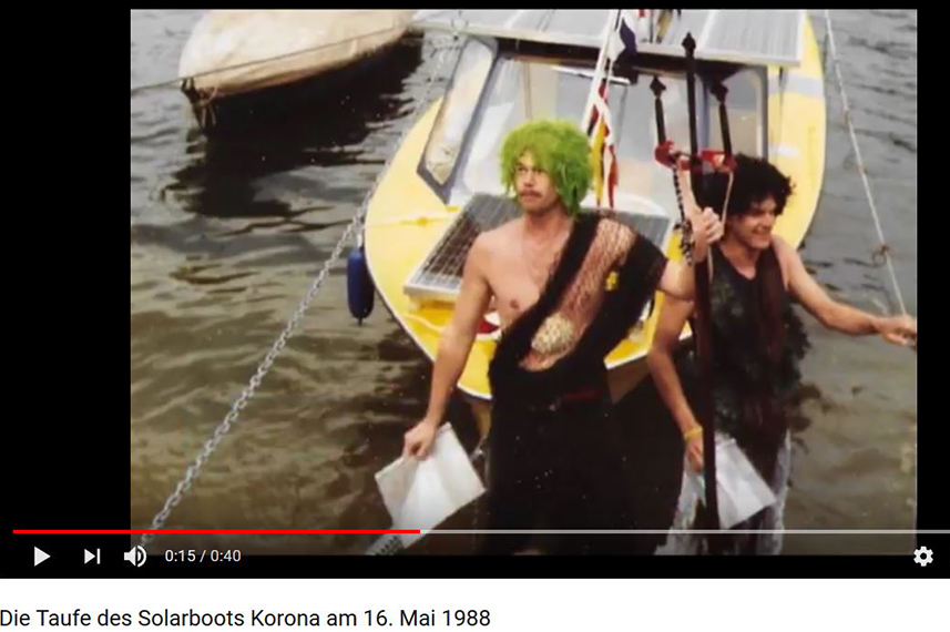 Screenshot aus dem YouTube-Video über die Taufe der Korona: Zwei als Wassermänner verkleidete Männer stehen im Wasser vor dem Boot. 