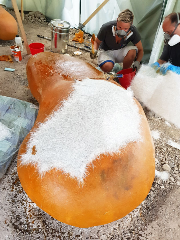 Zwei Männer mit Atemmaske arbeiten an einem Sitzelement. Darauf liegt eine Glasfasermatte.