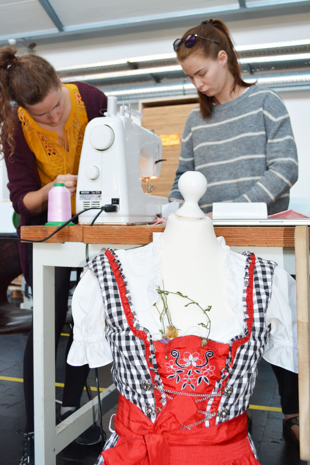 Zwei Frauen arbeiten an einer Nähmaschine. Im Vordergrund steht eine Schneiderpuppe. Sie trägt ein Dirndl.