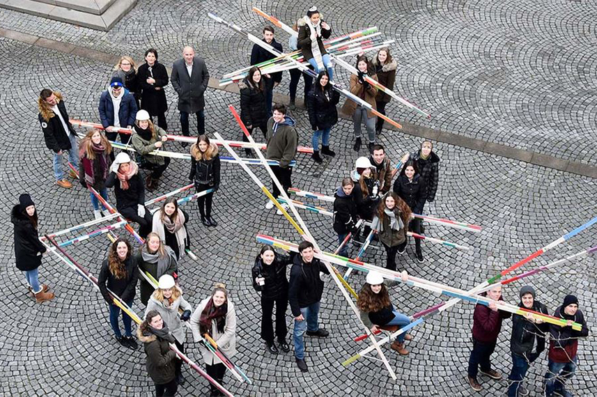 Luftaufnahme einiger Menschen, die auf einem gepflasterten Platz stehen und riesige Mikado-Stäbe auf den Schultern tragen oder in den Händen halten.