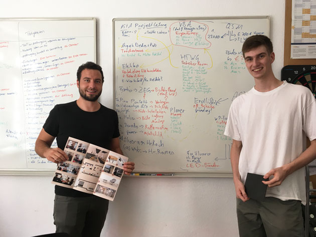 Zwei junge Männer vor einem vollgeschriebenen Whiteboard. einer der beiden hält einen geöffneten Prospekt in den Händen.