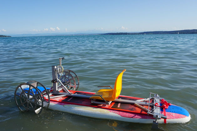 Ein Tretboot gebaut aus einem Surfbrett liegt am Seeufer im seichten Wasser.