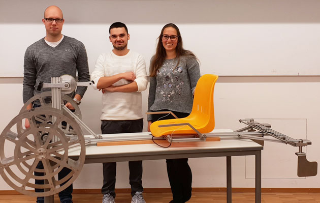 Drei Studierende stehen hinter einem Tisch. Auf ihm liegen Sitz und Fahrgestell ihres selbstgebauten Tretbootes.