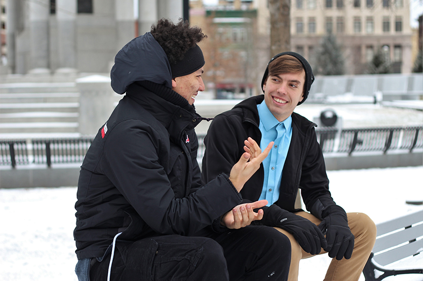 Zwei junge Männer in Winterkleidung sitzen auf der Lehne einer Parkbank und unterhalten sich.