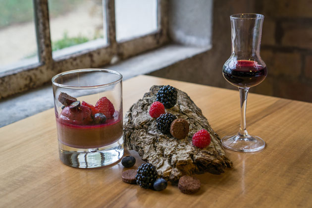 Auf einem Stück Baumrinde liegen Brombeeren und Himbeeren. Rechts daneben steht ein Grappa-Glas, zu einem Drittel gefüllt mit einer roten Flüssigkeit. Links der Rinde steht ein Glas zu einem Drittel gefüllt mit Schokoladen-Mousse, darauf liegen Brombeeren und Himbeeren sowie Schokoladenraspeln.