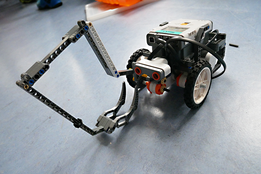 Ein vierrädriger Mind-Storm-Roboter mit einem langen Greifarm steht auf einer grauen Fläche.