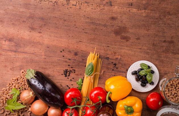 Blick aus der Vogelperspektive auf eine Holzoberfläche, auf der Paprika, Aubergine, Tomaten, Zwiebeln und Kräuter liegen.