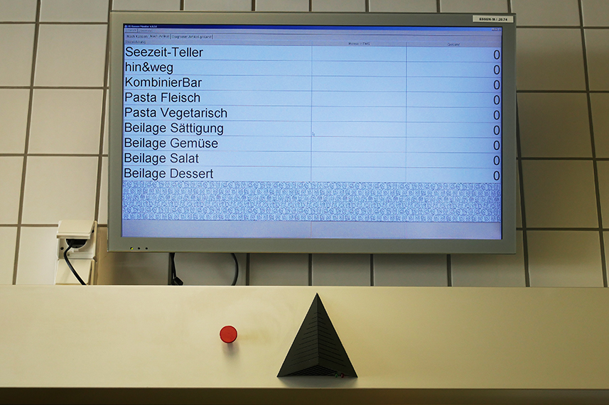 Zu sehen ist ein Bildschirm vor einer weiß gefliesten Wand. Auf dem Bildschirm stehen untereinander die Namen verschiedener Gerichte.