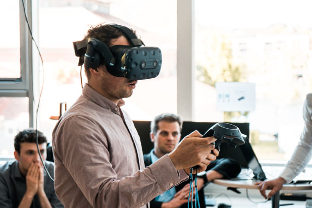 Zu sehen ist der Oberkörper eines jungen Mannes in einem rosa Hemd. Er trägt eine VR-Brill und hält einen Hand-3D-Scanner mit der rechten Hand hoch.