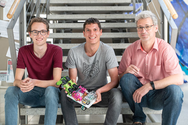 Die Informatik-Studenten Markus Käppeler (links) und Sebastian Rätzer (Mitte) sitzen mit Laboringenieur Jürgen Keppler (rechts) auf einer Treppe und blicken in die Kamera. Sebastian Rätzer hält einen bunten Krabbelroboter in seinen Händen.