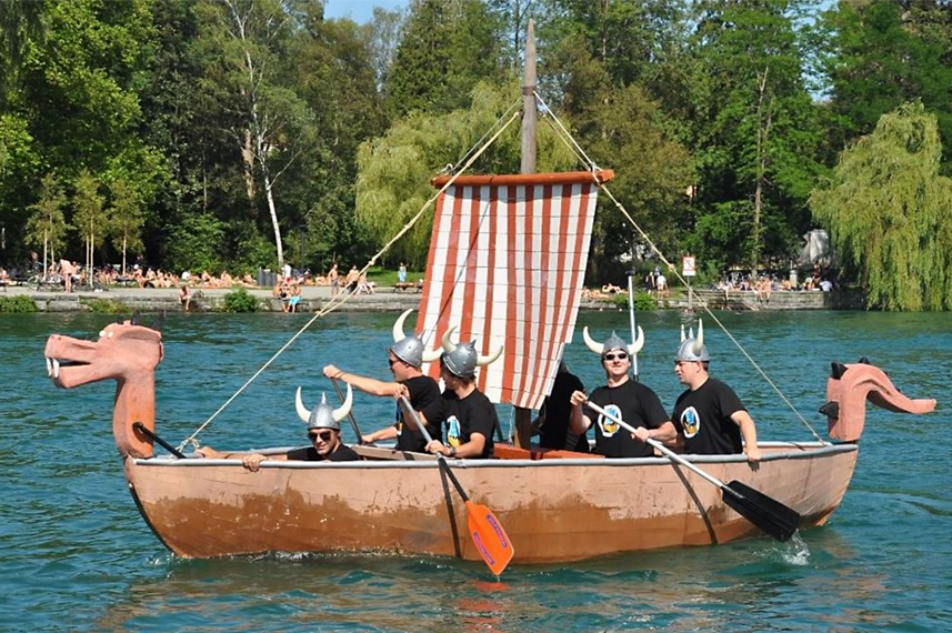 Fünf junge Männer in schwarzen T-Shirts und Wikinger-Helmen sitzen in einem Wikinger-Boot mit rot-weiß-gestreiftem Segel, das im Wasser fährt.