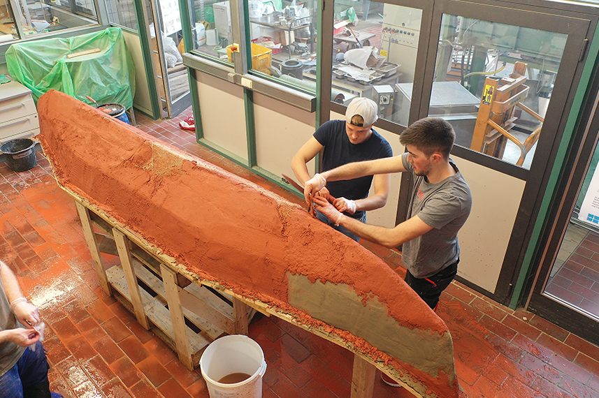 Blick von der Seite auf die Unterseite eines Kanus. Zwei Studenten arbeiten an dem Rumpf, der auf einem Holzständer aufliegt.