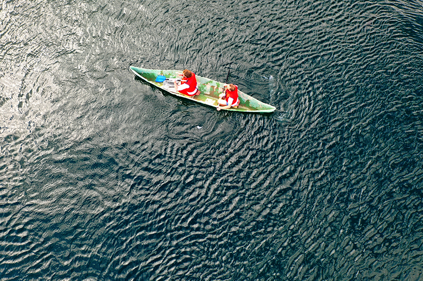 Blick aus der Vogelperspektive auf ein grünes Kanu mit zwei rot gekleideten Insassen. Das Kanu fährt im Wasser.