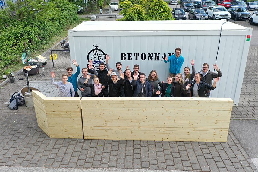 Eine Gruppe von zirka 15 Studierenden steht zwischen einer Holzwand und einem Container mit der Aufschrift "Betonkanu"