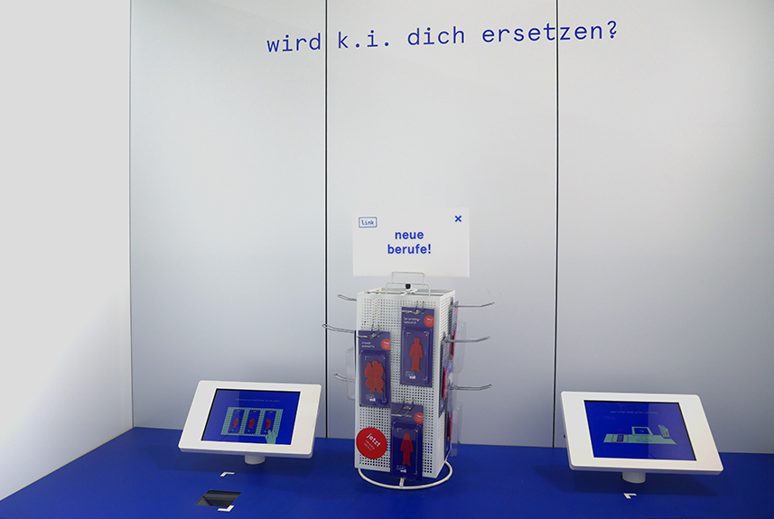 Blick in einen Ausstellungsraum. An der weißen Wand steht in blauen Buchtstaben: Wird KI Dich ersetzen? Auf einem blauen Tisch sind zwei Tablets angebracht, daneben steht ein Ständer mit Kärtchen, auf denen unterschiedliche Berufsbilder und ihre Zukunft in der digitalen Transformation beschrieben sind.