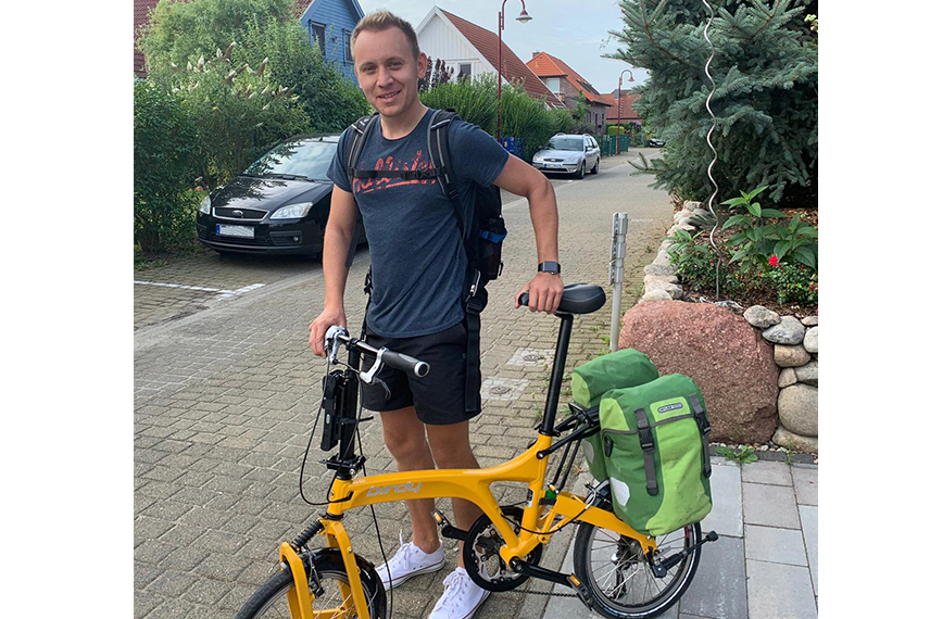 Max Pabst steht links neben einem orange-gelben Faltrad, auf dem grüne Fahrradtaschen angebracht sind.