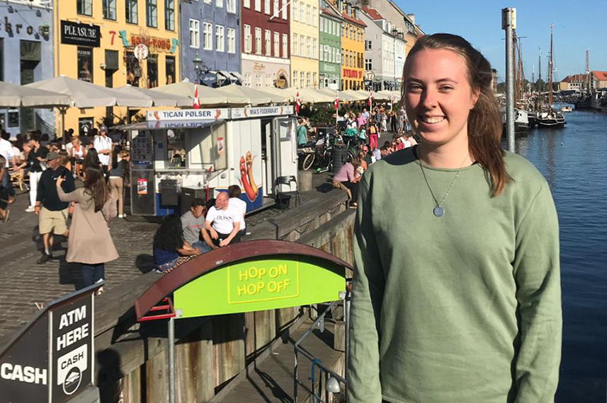 Ann-Kathrin Straßer steht auf einer Brücke in Kopenhagen. Rechts hinter ihr ist Wasser zu sehen, links leuchten die bunten Fassaden von Häusern.