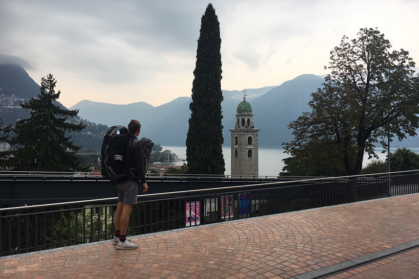 Zu sehen ist Florian Jungheim von hinten. Er blickt von einem Gehweg hinunter auf den Lago Maggiore, hinter der Wasseroberfläche ragen Berge auf.