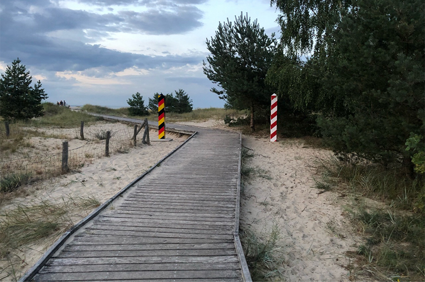 Ein Holzsteg führt durch Dünen. Links davon steht ein schwarz-rot-gold-gestreifter Pfosten, rechts ein rot-weiß-gestreifter Pfosten.