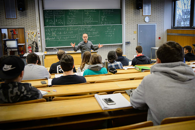 Prof. Dr. Bernd Jödicke steht an der Tafel eines Hörsaals und breitet die Arme aus. Vor ihm sitzen in Holzbänken Studierende.