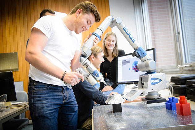 Ein Student führt den Arm eines Mehrachsroboters zu einem grauen Gegenstand auf einer grauen Platte. Im Hintergrund arbeiten Studierende am Bildschirm.