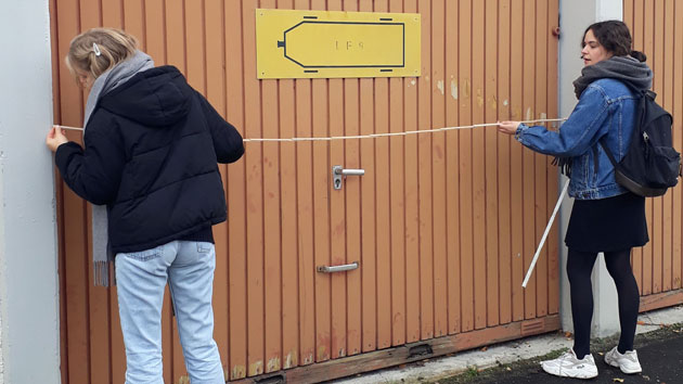 Zwei Studentinnen stehen rechts und link von einem Garagentor und strecken gemeinsam ein Maßband über die Breite des Tores.