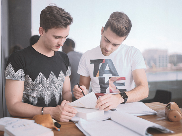 Zwei Studenten stehen an einem Schreibtisch und blicken gemeinsam in ihre Aufzeichnungen, die in Blättern auf dem SChreibtisch liegen.