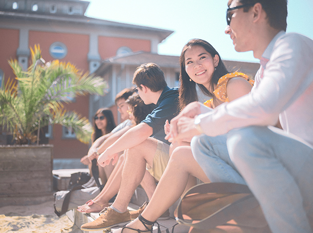 Seitenansicht auf sechs Studentinnen, die in einer Reihe nebeneinander am Rand eines Sandkastens der Strandbar der Hochschule sitzen.