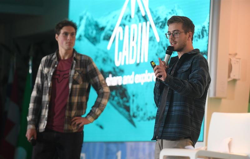 Michael Hendlich (links) und Finn Wilkesmann (rechts) stehen vor einer Leinwand, auf der das „MyCabin“-Logo vor schneebedeckten Bergen zu sehen ist. 